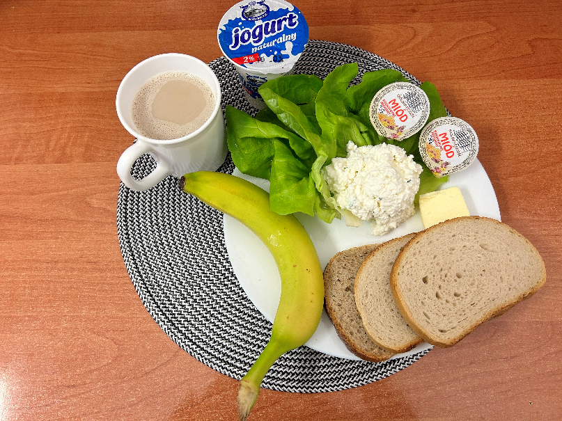 Na zdjęciu znajduje się: Jogurt naturalny, Kawa zbożowa z mlekiem, Chleb mieszany pszenno-żytni, Chleb Graham, Masło extra 82%, Twarożek, Miód, Banan, ​​​​​​​Sałata zielona